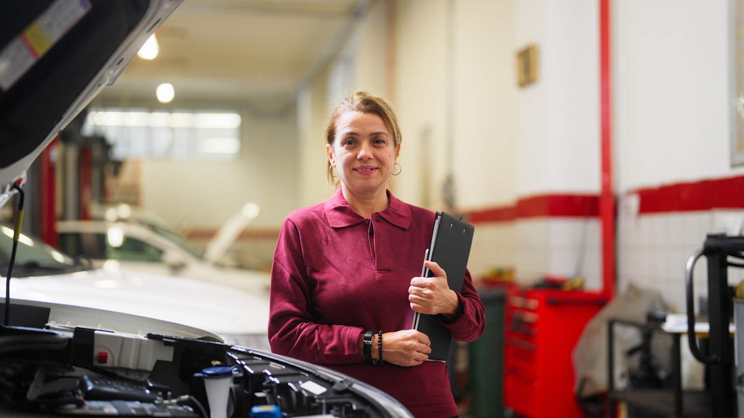 woman in red blouse with notebook standing next to car with hood lifted